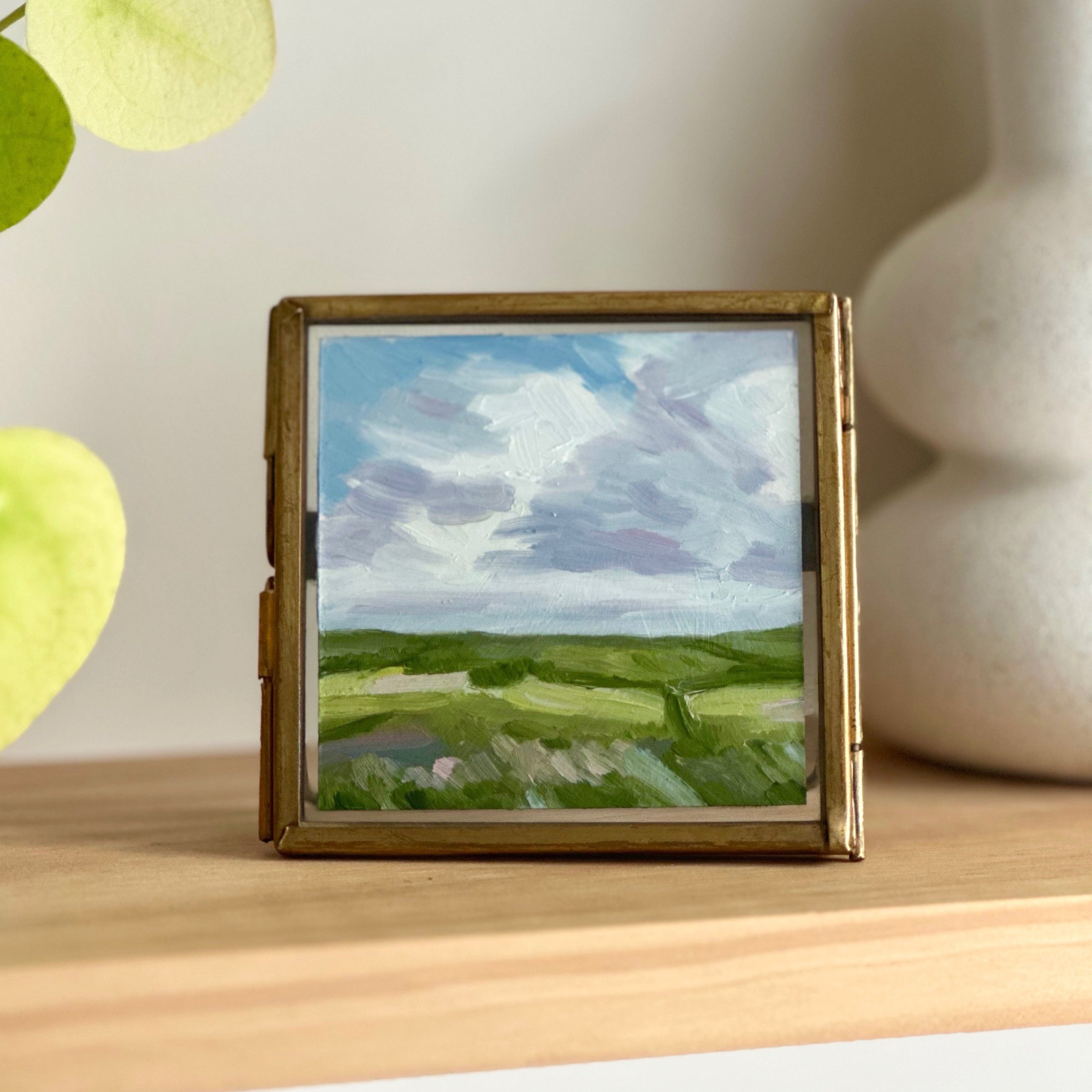 Small framed painting of a landscape on a wooden shelf with a vase and leaves in the background.