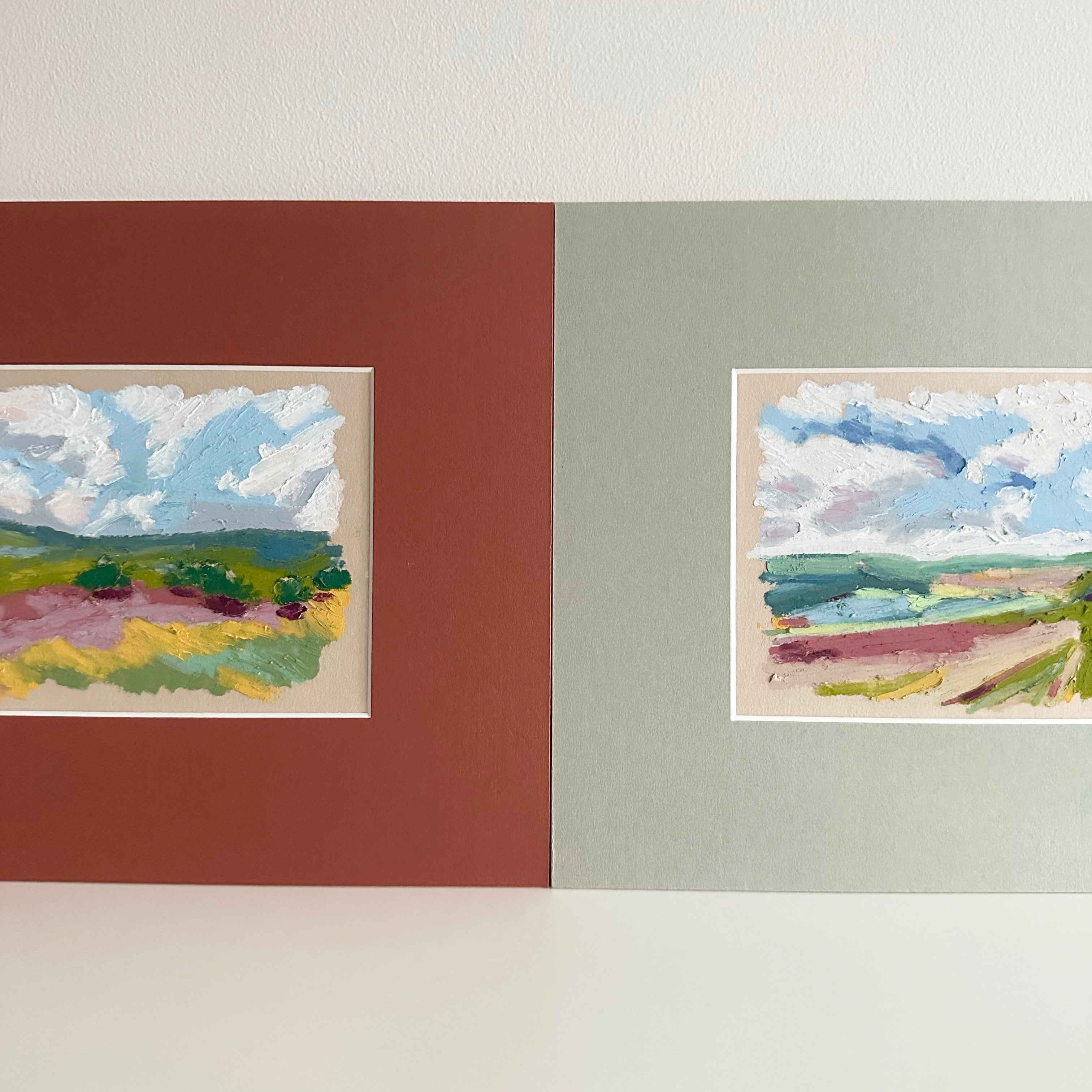 Two landscape paintings displayed on a wall with a terracotta red and green mounts.