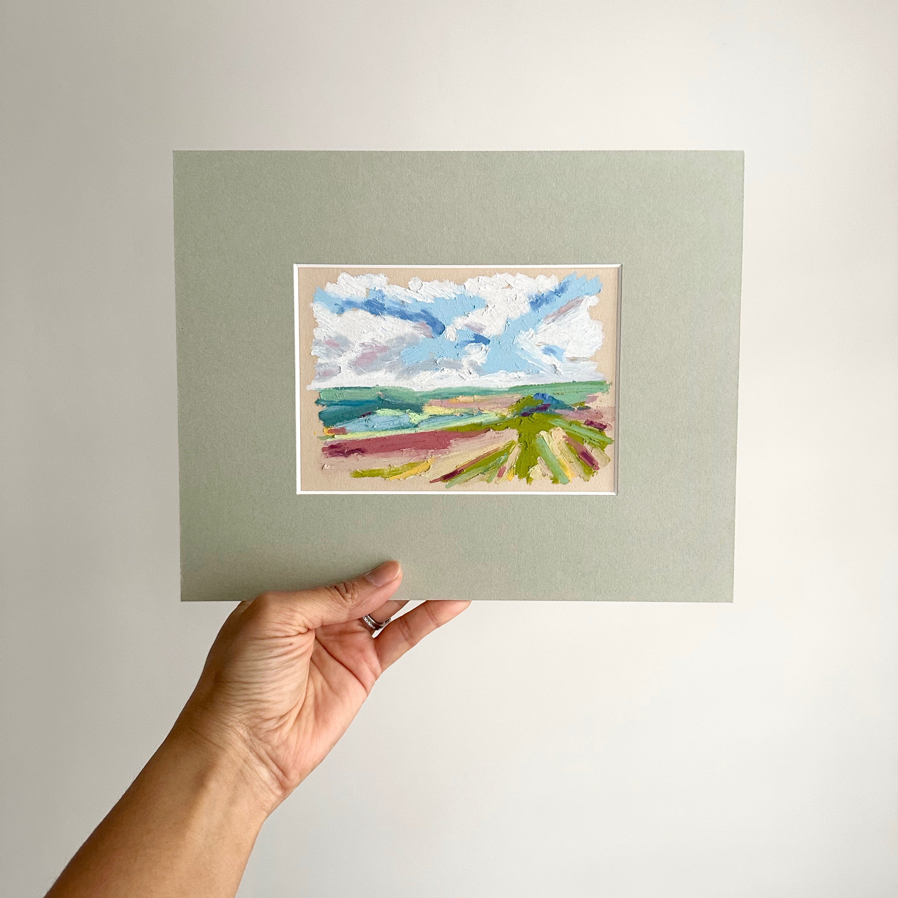 Hand holding a small landscape painting with a neutral background