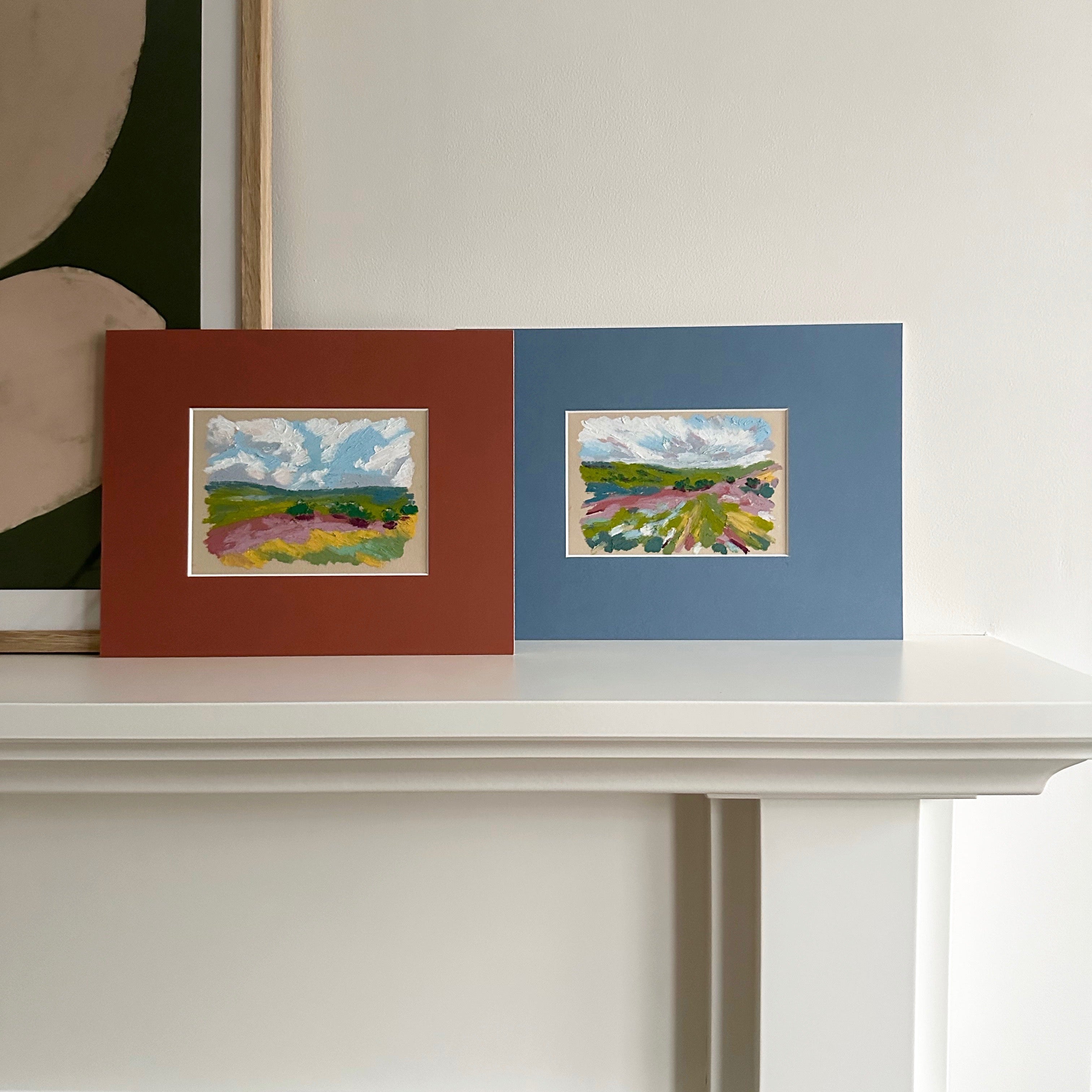 Two framed landscape artworks on a white mantel piece.
