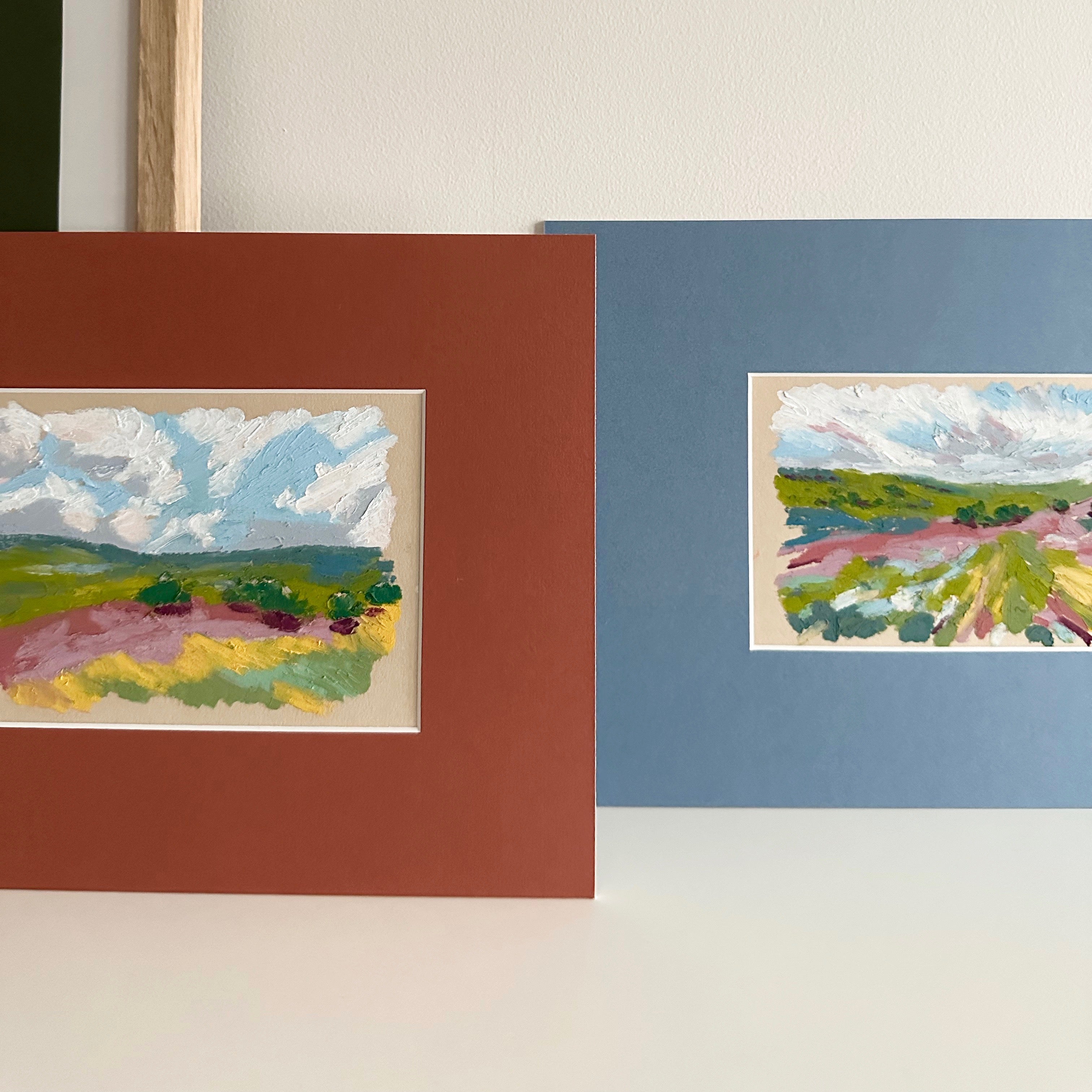 Two landscape paintings in matted frames on a white surface
